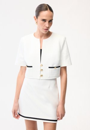 Vrouw met naar achteren gestyled haar, gekleed in een witte cropped blazer met gouden knopen en een bijpassende minirok met zwarte bies.