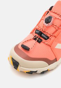 Zapatilla deportiva naranja y rosa con tejido texturizado, cierre de cordones negro y una caperuza de goma marrón en la puntera. Acentos notables en la suela en amarillo y negro.