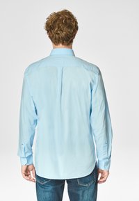 Camicia azzurra chiara a maniche lunghe con orlo dritto, piega centrale posteriore singola e polsini con bottone, realizzata in tessuto morbido, indossata con jeans scuri.