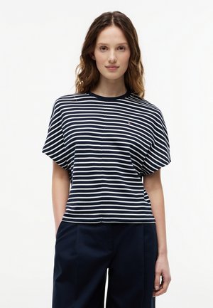 Donna con capelli ondulati che indossa una maglietta a maniche corte a righe blu navy e bianco e pantaloni blu navy scuro, in piedi contro uno sfondo bianco semplice.
