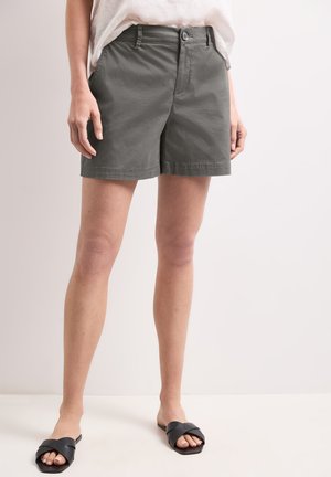 Person iført grå knælang shorts, en hvid top og sorte åbne slip-on sandaler, stående mod en lys baggrund.
