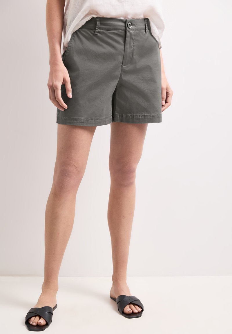 Persona che indossa pantaloncini grigi al ginocchio, una maglietta bianca e sandali neri aperti senza cinturini, in piedi su uno sfondo chiaro.