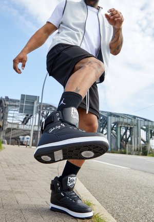Homme en short noir et débardeur blanc avançant, portant des baskets montantes noires avec semelles blanches sur un trottoir en ville près d’un pont métallique.