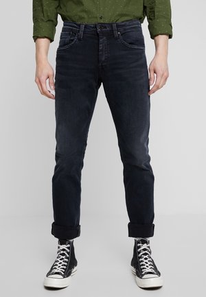 Mand iført mørke rullede jeans, sorte høje sneakers med hvide snørebånd og en grøn skjorte med knapper med opkrogede ærmer.