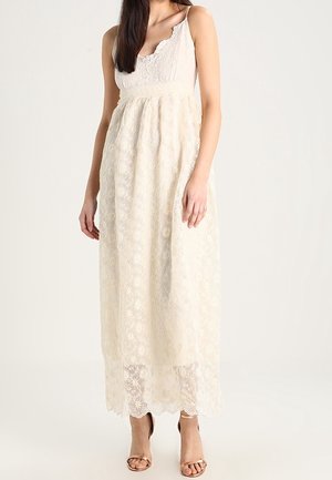 Frau trägt ärmelloses weißes Maxi-Kleid aus Spitze mit Blumenmuster und geschwungenem Ausschnitt, kombiniert mit offenen Absätzen, stehend vor einfarbigem Hintergrund.