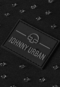 Czarna, teksturowana tkanina z naszywką z logo "JOHNNY URBAN" w wytłoczonych literach, z małymi, przezroczystymi kroplami rozsianymi po powierzchni.
