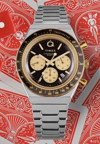 Die Timex Q Chronograph-Uhr verfügt über ein schwarzes Zifferblatt mit goldenen Akzenten, goldenen Zusatzzifferblättern, Tachymeter, einem silbernen Edelstahlarmband und einem rechteckigen Gehäuse.