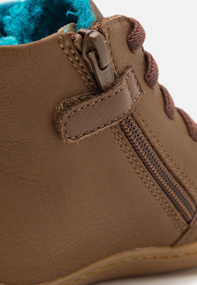 Botín de cuero marrón hasta el tobillo con cremallera lateral, cordones marrones y un forro de felpa azul brillante, con material texturizado y costuras resistentes.