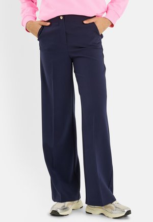 Persona con pantalones anchos azul marino, sudadera rosa claro, manos en los bolsillos y zapatillas deportivas blancas de pie contra un fondo sencillo.