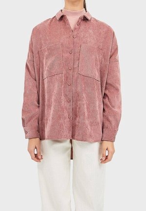 Woman wearing a loose dusty pink button-up corduroy shirt with two chest pockets and cream-colored pants.