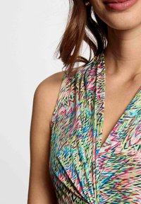 Bunt gemustertes ärmelloses Kleid mit einem drapierten Ausschnitt, dekoriert mit Zickzackgrafiken in Rosa-, Grün- und Blautönen auf einem hellen Hintergrund.