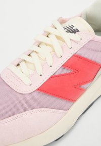 Baskets rose clair en mesh et daim. Elles présentent un logo rouge vif, des lacets crème et une semelle texturée à finition lisse.