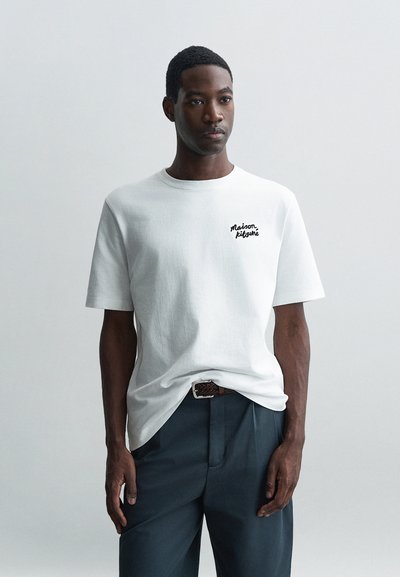 T-shirt en coton blanc avec un col rond, arborant un petit logo noir brodé sur la poitrine gauche. Porté avec un pantalon sombre ajusté.