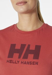Camiseta de algodón roja con un escote redondo que presenta un logo "HH" en un marrón oscuro y la frase "HELLY HANSEN" en letras bordadas y en negrita.