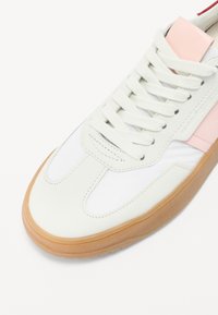 Beli in roza športni copat z gladkim usnjenim zgornjim delom, vezalkami in gumijastim podplatom z teksturiranimi poudarki.