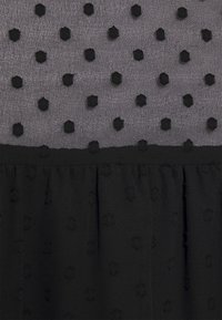 Svart klänning med genomskinlig grå överdel som har upphöjda svarta prickmönster; den nedre delen är plisserad. Strukturerad tyg med ett lagerdesign.