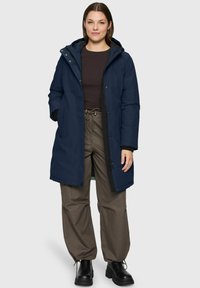 Woman wearing a navy hooded winter coat, black long-sleeve shirt, olive drawstring pants, and black boots, standing against a light background.
