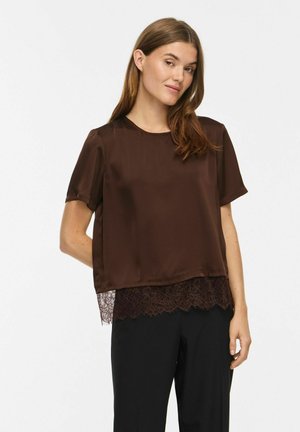 Femme portant un chemisier en satin marron à manches courtes avec une bordure en dentelle à l'ourlet, associé à un pantalon noir, debout devant un fond blanc.