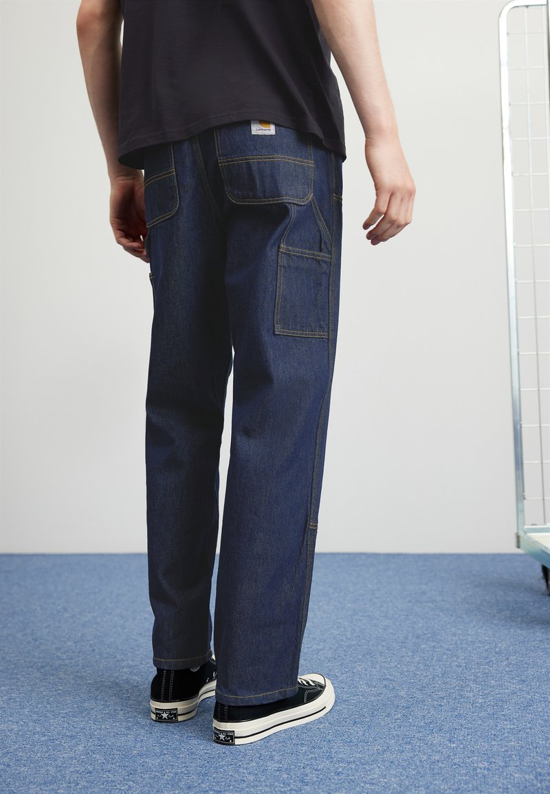 Dark blue denim trousers with a relaxed fit, featuring double pockets and contrast stitching, paired with black sneakers.