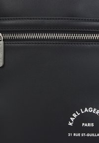 Čierna kožená taška s textúrovaným povrchom, ktorá má výrazný strieborný zips, minimalistický dizajn loga a biele písmená.