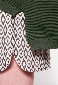 Grön ribbad stickad tröja överlappar en mönstrad kjol med svarta diamantformer på en krämfärgad bakgrund och en böjd nederkant.