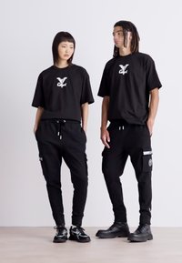 Twee modellen dragen bijpassende zwarte oversized t-shirts met een wit logo, gecombineerd met zwarte joggers met zakken en accenten.