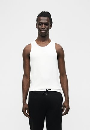 Młody czarnoskóry mężczyzna z krótkimi dredami, ubrany w biały bezrękawnik i czarne spodnie z wiązaniem, stojący na jednolitym tle.