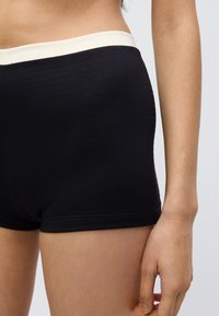 Svarte ribbestrikkede shorts med en kremfarget elastisk linning, med en tettsittende design og myk tekstur. Nærbilde som viser en hånd på låret.