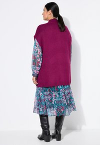 Magenta Strickweste mit hohem Kragen, über einem türkisfarbenen Blumenkleid mit Rüschensaum getragen. Schwarze kniehohe Stiefel runden das Outfit ab.