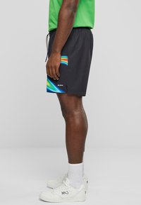 Svarte badshorts med regnbuegradientdesign på venstre side, med elastisk midje og snøring. Hvite sneakers med logo.