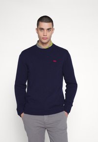 Maglione lavorato a maglia blu navy con collo rotondo, polsini e orlo a coste, con un piccolo logo rosso sul petto e indossato sopra una camicia con colletto.