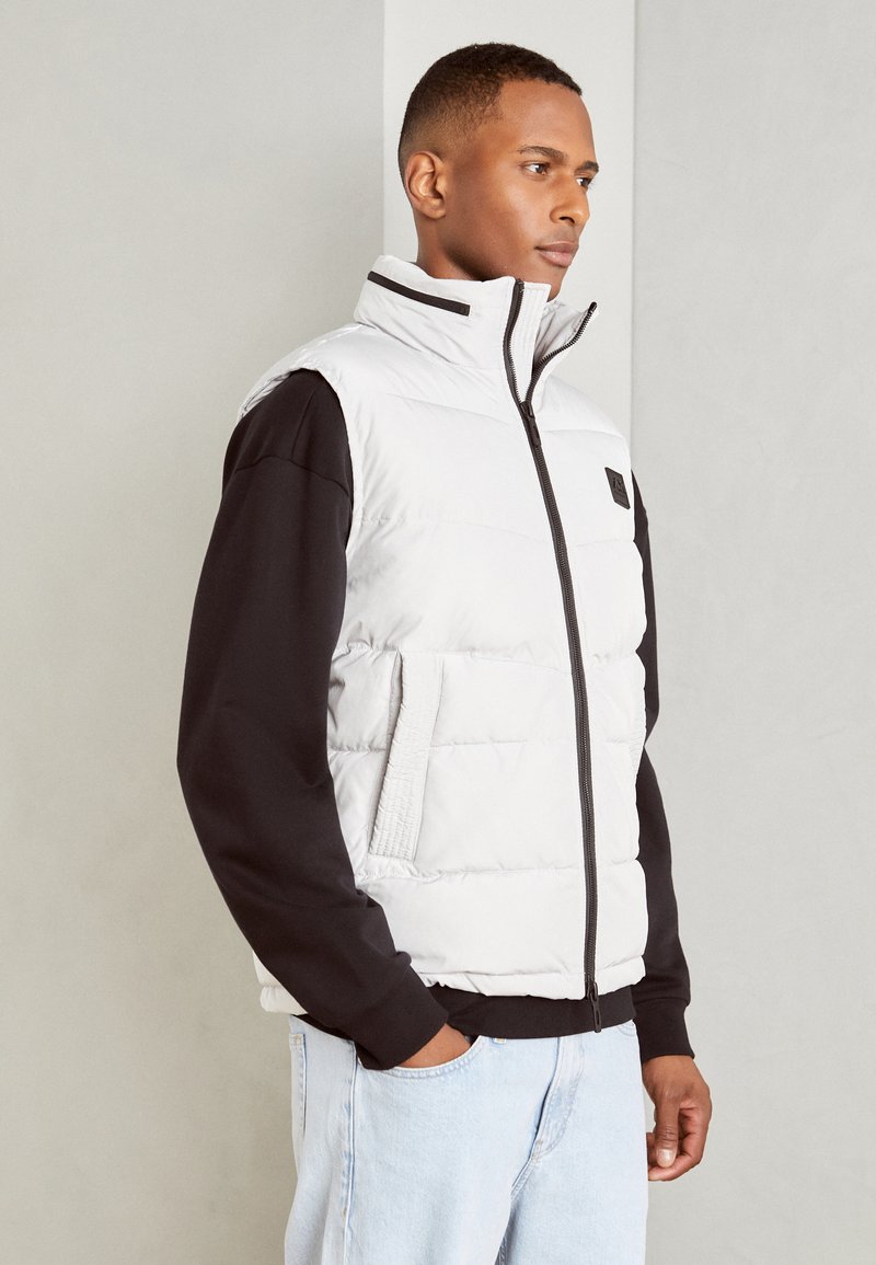 Gilet rembourré blanc avec col montant, fermeture éclair à l'avant, poches latérales, et un patch logo noir, porté sur une chemise noire à manches longues et un jean clair.