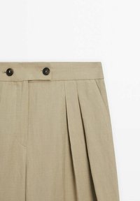 Pantalon plissé beige avec une taille haute ornée de deux boutons foncés et des poches latérales sur fond blanc.