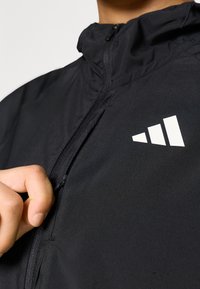 Chaqueta deportiva negra con cuello alto, cremallera frontal y un logo de rayas blancas en el lado derecho. El material parece ser ligero y suave.
