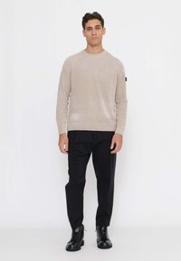 Hellbeiger Strickpullover mit gerippten Schulterdetails, kombiniert mit schwarzen Hosen und schwarzen Schnürstiefeln, stehend vor weißem Hintergrund.