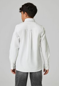 Witte lange mouwen shirt met een standaard kraag, met een rugplooi en knoopmanchetten, gedragen met grijze broeken. Gladde stoftextuur.