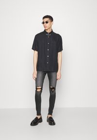 Camisa preta de manga curta com botões e bolso, combinada com jeans skinny cinzas rasgados e mocassins pretos. Os acessórios incluem óculos de sol escuros.