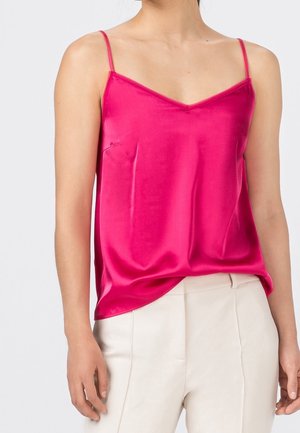 Frau trägt ein hellrosa Satin-Camisole mit dünnen Trägern und cremefarbene hochtaillierte Hose vor einfarbigem Hintergrund.