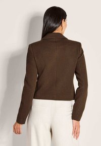 Vrouw met lang donker haar draagt een donkerbruin textuurblazer en lichtbeige broek, staand met haar rug naar de camera tegen een effen lichte achtergrond.