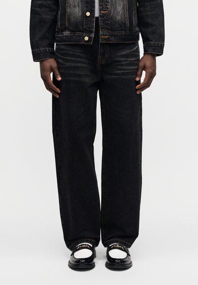 Μαύρο τζιν μπουφάν και ταιριαστό μαύρο, φαρδύ τζιν παντελόνι με κίτρινο ράψιμο και χρυσά μεταλλικά στοιχεία, σε συνδυασμό με μαύρα παπούτσια.