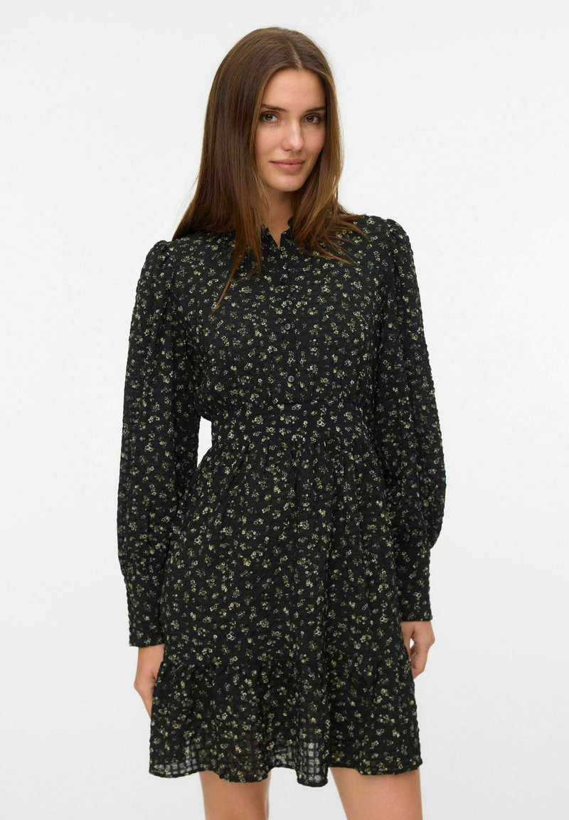 Zwarte jurk met een bloemenpatroon in groen en wit, lange poffcuffs, een knoopsluiting aan de voorkant en een uitlopende zoom.