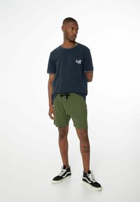T-shirt bleu marine à manches courtes avec poche poitrine, associé à un short vert et des baskets montantes noires. Présente des surpiqûres contrastantes sur le t-shirt.