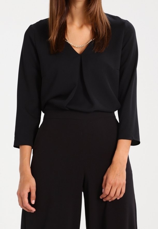 Femme portant un chemisier noir à manches longues avec un col en V et un pantalon noir taille haute, avec un collier chaîne en or visible au niveau de l'encolure.