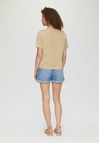 Beige Baumwoll-T-Shirt mit lässiger Passform, kombiniert mit hellblauen Jeansshorts, die mit umgeschlagenen Säumen und braunen Sandalen abgerundet sind.