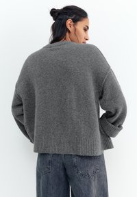 Žena s tmavými vlasy staženými do drdolu, oblečená v volném šedém pleteném svetru a vybledlých modrých džínách, stojící bokem na bílém pozadí.