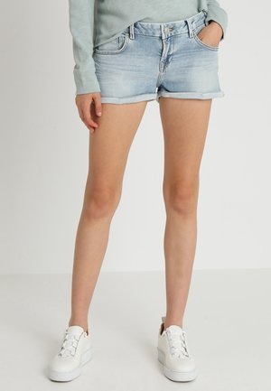 Persona que lleva pantalones cortos de mezclilla azul claro, camiseta de manga larga gris y zapatillas blancas con plataforma, de pie contra un fondo liso.