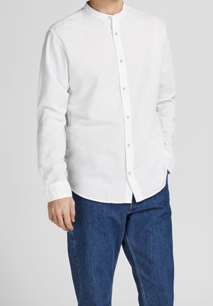 Mężczyzna ubrany w białą koszulę zapinaną na guziki z kołnierzykiem typu mandarynka i niebieskie dżinsy, stoi z jedną ręką w kieszeni na jednolitym tle.