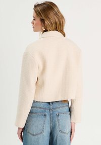 Gekürzte Jacke aus einem strukturierten, hellbeigen Stoff mit langen Ärmeln und einem einfachen Kragen, kombiniert mit hochgeschnittenen blauen Jeans.