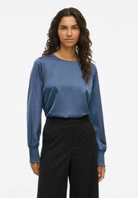 Vrouw met lang, krullend haar, gekleed in een blauw satijnen blouse met smockmanchetten en hoge, zwarte broeken, staand tegen een effen achtergrond.