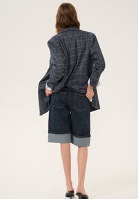 Blazer a cuadros en azul marino con hombros estructurados, combinado con pantalones cortos de mezclilla sueltos con dobladillo doblado y bolsillos traseros. Zapatos negros tipo slip-on.